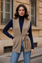 Camel merino wool belted sweater vest with V-neck and waist tie, worn over a navy turtleneck.